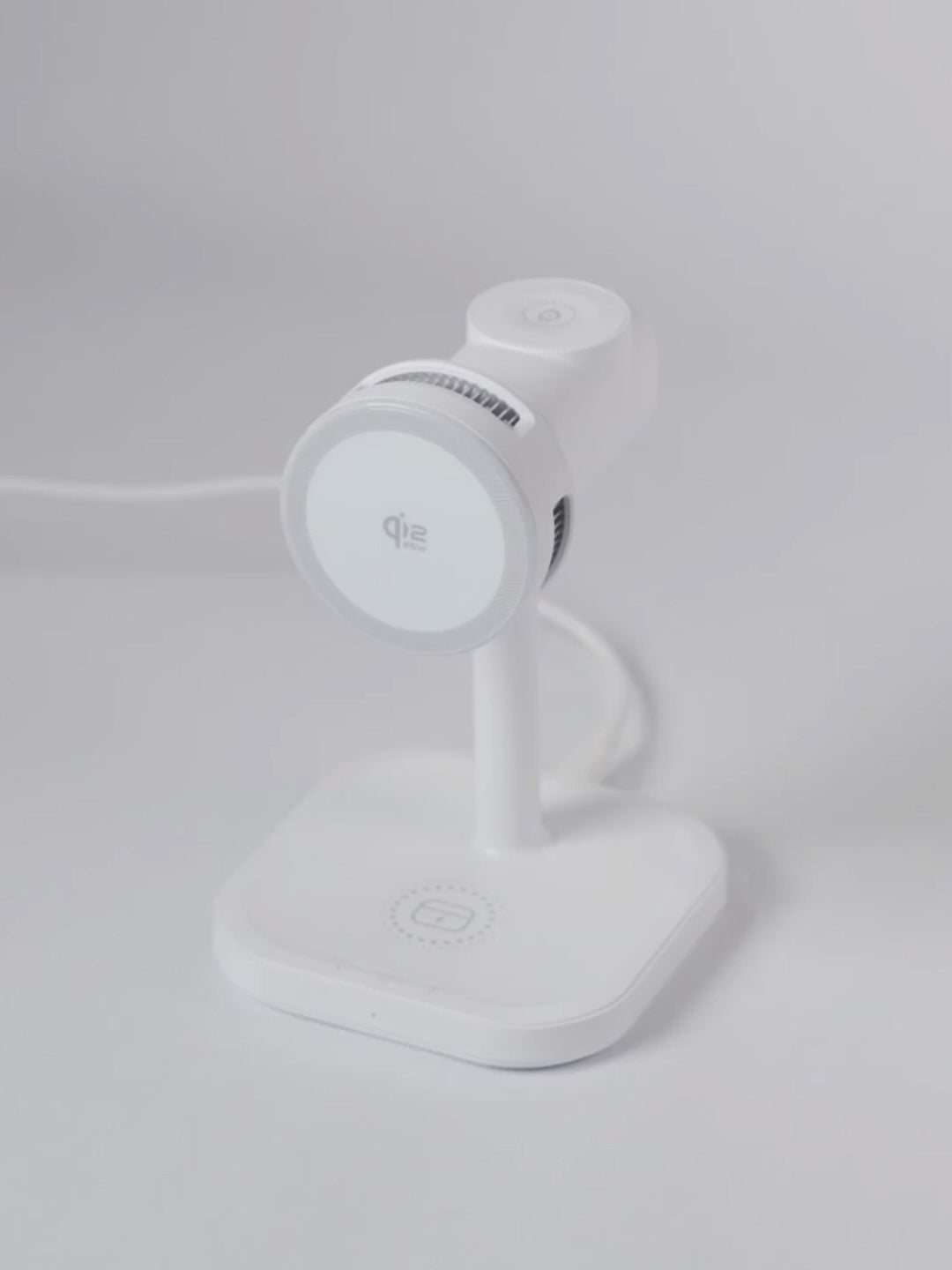 A demonstration video of the white Schlüssel 3-in-1 wireless charging station. A hand sequentially places an Apple Watch on the top dock, AirPods on the base, and an iPhone horizontally on the front magnetic Qi2 pad, showing all three devices charging simultaneously