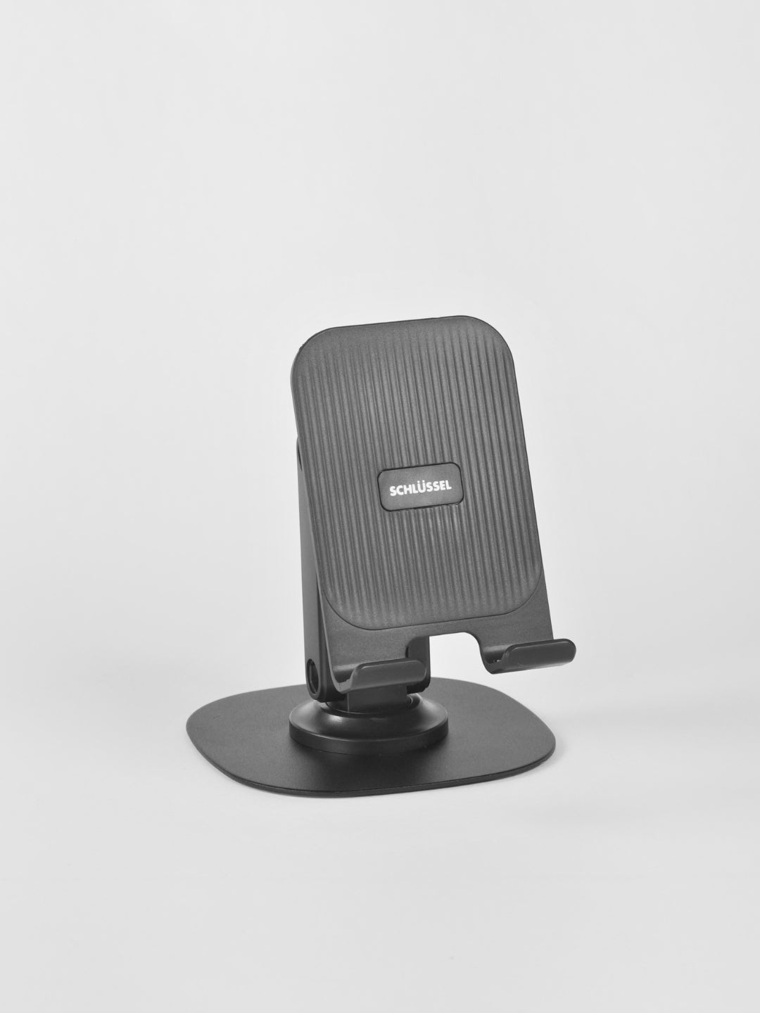 A standalone black Schlüssel adjustable desktop phone stand featuring a vertically ridged backrest and a wide, flat base plate, captured from a three-quarter angle against a neutral gray background